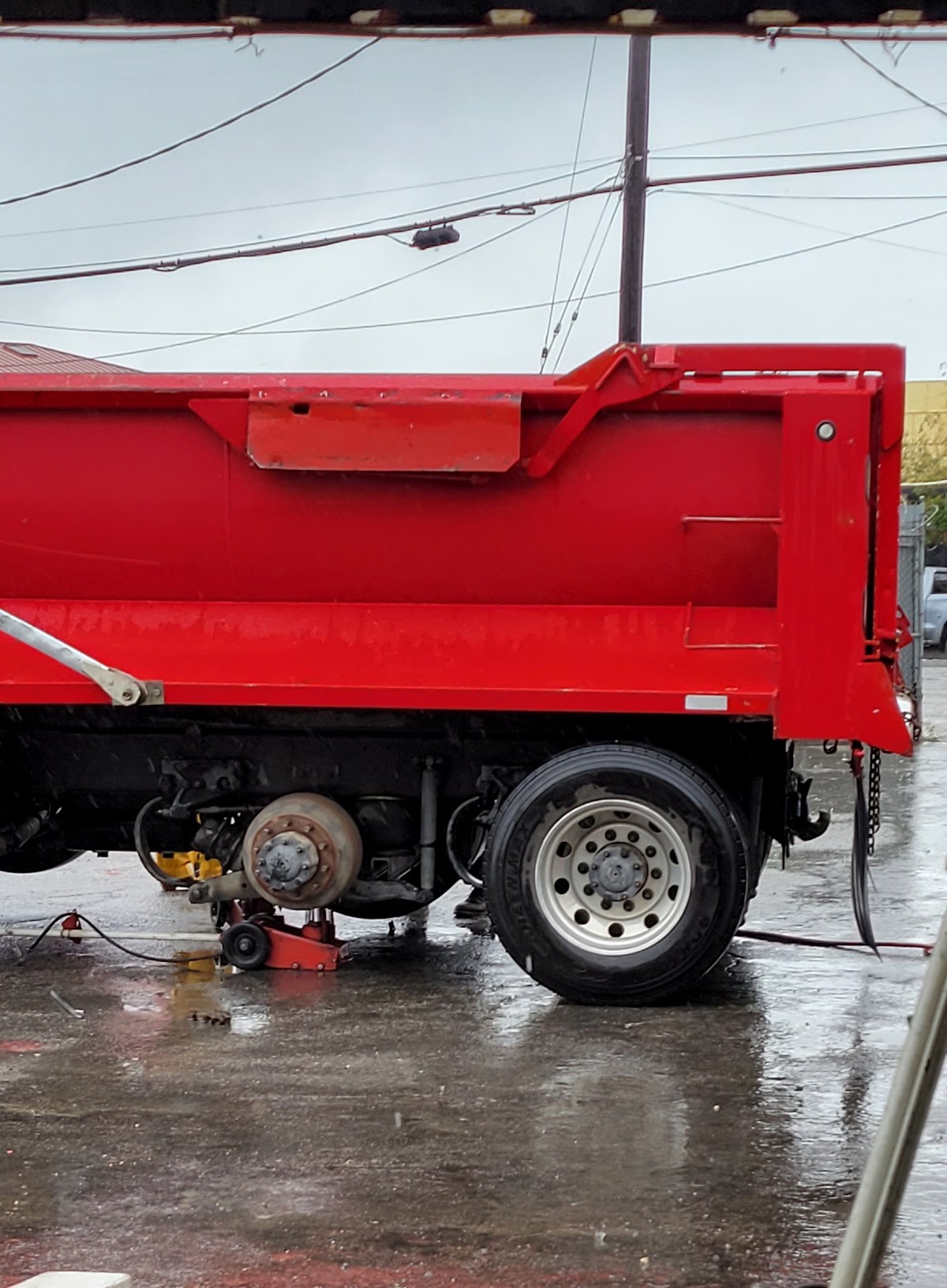 A red dump truck with a wheel removed and a jack under the axle, in the rain